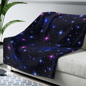 Puede incluir: Una manta suave y mullida con un fondo azul oscuro y un patr&oacute;n repetitivo de estrellas blancas, azules y rosas. La manta est&aacute; drapeada sobre un sof&aacute; blanco.