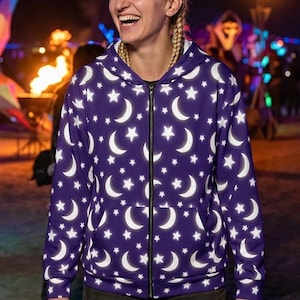 May include: A long-sleeve zip-up hoodie featuring a repeating pattern of white crescent moons and stars against a deep purple background. The garment includes a hood, a full-length front zipper, and two front pockets, worn over dark cargo pants.