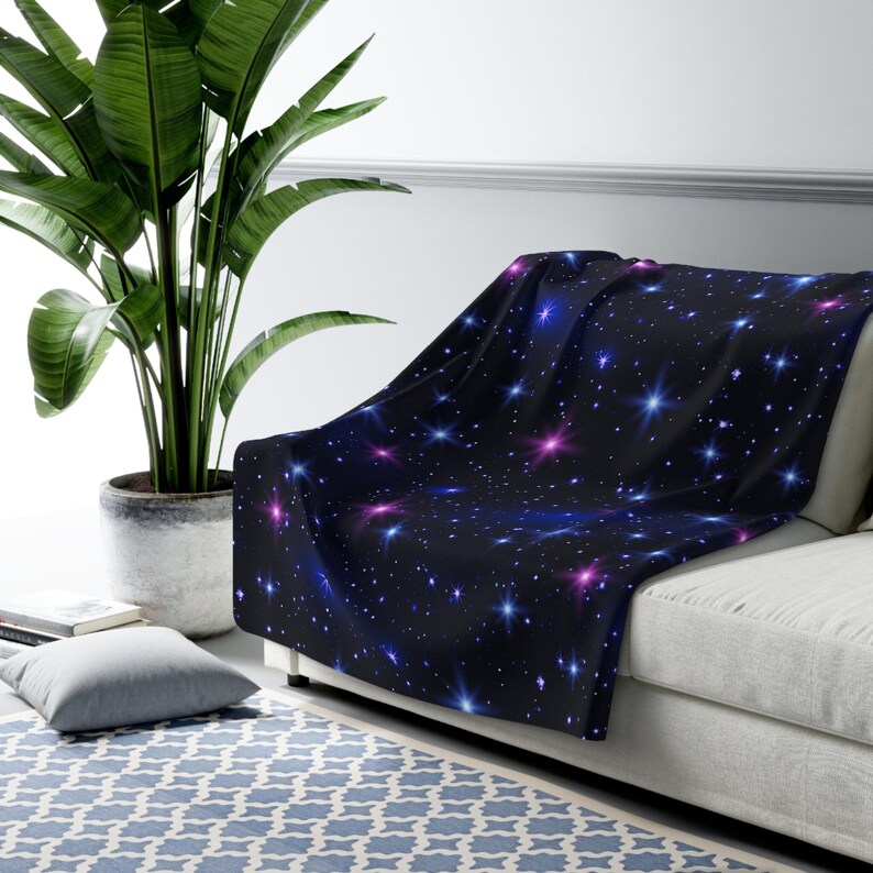 Puede incluir: Una manta suave y mullida con un fondo azul oscuro y un patr&oacute;n repetitivo de estrellas blancas, azules y rosas. La manta est&aacute; drapeada sobre un sof&aacute; blanco.