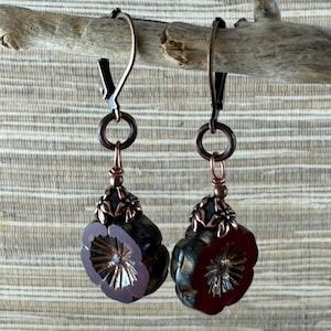 May include: A pair of handcrafted earrings featuring dark red flower-shaped pendants with intricate details. The earrings have copper-colored metal accents and are suspended from a simple hook. The background is a neutral, textured surface.