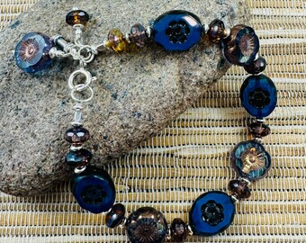 May include: A handcrafted bracelet featuring a mix of dark blue, brown, and amber-colored beads. The bracelet includes oval and round beads with floral designs, connected by a silver-toned chain. The bracelet is displayed on a gray stone.