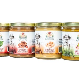 May include: Four glass jars of nut butter with gold lids. The labels read "Hazelnut Silky Butter", "Pecan Silky Butter", "Cashew Silky Butter", and "Pistachio Silky Butter". Each jar features a smiling man in a hat and overalls.