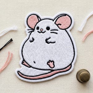 May include: Embroidered patch featuring a cute, cartoon-style white mouse with pink ears and a pink tail. The mouse is smiling and has black eyes and whiskers. The patch is outlined in black and is on a cream-colored fabric.