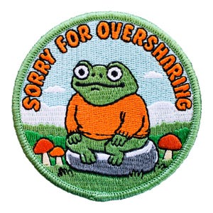 May include: Embroidered patch with a green frog wearing an orange sweater, sitting on a rock. The patch has a light blue background with the text "SORRY FOR OVERSHARING" in orange. The patch is round with a green border.