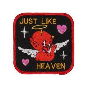 May include: Embroidered patch with a red devil character with wings, halo, and horns. The patch is black with the words "JUST LIKE HEAVEN" in yellow. Two pink hearts and two white crosses complete the design. The patch has a red border.