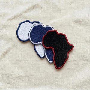 May include: Three embroidered patches in the shape of the African continent. One is white with a navy blue border, another is navy blue with a white border, and the third is black with a red border. The patches are arranged on a beige fabric.