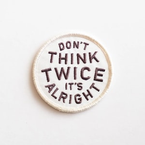 May include: A circular embroidered patch with the words "DON'T THINK TWICE IT'S ALRIGHT" in dark brown letters. The patch is white with a cream-colored stitched border. The text is centered on the patch.