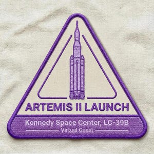 May include: Embroidered patch in a triangular shape with a purple border. The patch features a rocket illustration inside a white triangle, with the text "ARTEMIS II LAUNCH" and "Kennedy Space Center, LC-39B - Virtual Guest" in purple.