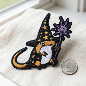 May include: Embroidered patch featuring a gecko wizard wearing a black hat with gold stars and a matching robe. The gecko holds a purple staff. The patch has a white belly and a yellow and black tail. A quarter is next to the patch for size reference.