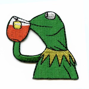 May include: Embroidered patch featuring a green frog drinking from a glass of tea. The frog has a yellow collar and a white eye. The tea is orange with a yellow tea bag. The patch is on a white background.