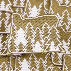 May include: Olive green embroidered patch in the shape of the state of Oregon. The patch features a white tree design filling the state's outline. The patch has a white border and a textured appearance.