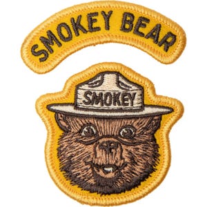 May include: Embroidered patch featuring Smokey Bear, a brown bear wearing a ranger hat. The patch has a yellow border and includes the text "SMOKEY BEAR" above the bear's head and "SMOKEY" on the hat.