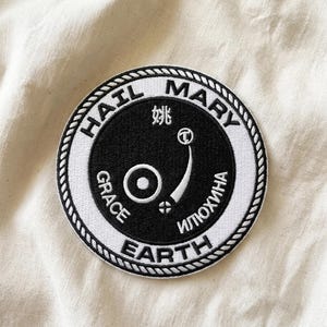 May include: Circular patch with a black center and white border. The text "HAIL MARY GRACE EARTH" is in a circular pattern around the edge. The center features a stylized design and text in multiple languages.