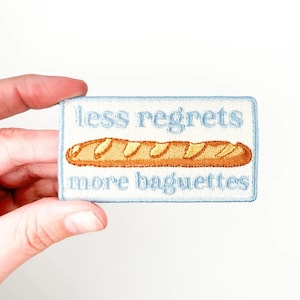 May include: Embroidered patch with a light blue border and the text "less regrets more baguettes" in light blue. A brown baguette is embroidered in the center. The patch is rectangular.