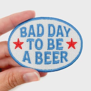 May include: Oval-shaped embroidered patch with the text "BAD DAY TO BE A BEER" in blue, surrounded by a blue border. Two red stars flank the text. The patch is held by a hand.