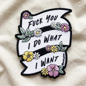 May include: Embroidered patch with the text "FUCK YOU I DO WHAT I WANT" on a white banner with black stripes. The patch is decorated with embroidered flowers in yellow, pink, and white, and green leaves.