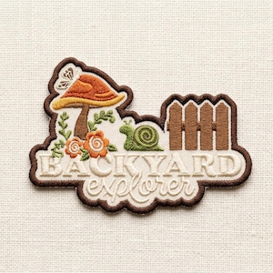 May include: Embroidered patch with a brown border featuring a mushroom, flowers, a snail, and a fence. The patch has the words "BACKYARD explorer" in a whimsical font. The mushroom has an orange cap and a butterfly.