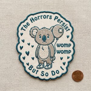 May include: Embroidered patch featuring a sad koala holding a balloon. The patch has the text "The Horrors Persist But So Do I" and "womp womp". The patch is surrounded by a teal border and teal hearts.
