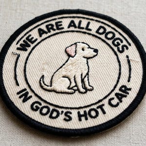 May include: Circular embroidered patch with a black border and the text "WE ARE ALL DOGS IN GOD'S HOT CAR". The patch features a cream-colored background with a seated cartoon dog illustration.