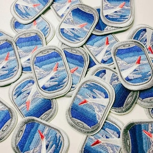 May include: A collection of embroidered patches shaped like airplane windows. Each patch features a view of a plane wing with a red tip against a blue sky and clouds. The patches have a gray border.