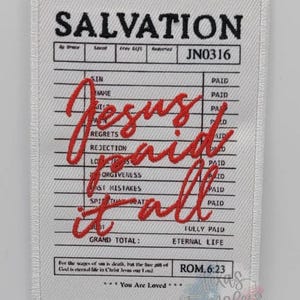 May include: A white rectangular patch with black text that reads "SALVATION" at the top. Red cursive text in the center says "Jesus Paid it All." The patch has a ledger-like design with various terms and the word "PAID" next to them.