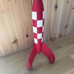 May include: A red and white rocket model with a checkered pattern on the body and three curved fins. The rocket has a pointed top and is standing on a wooden surface. A decorative item.