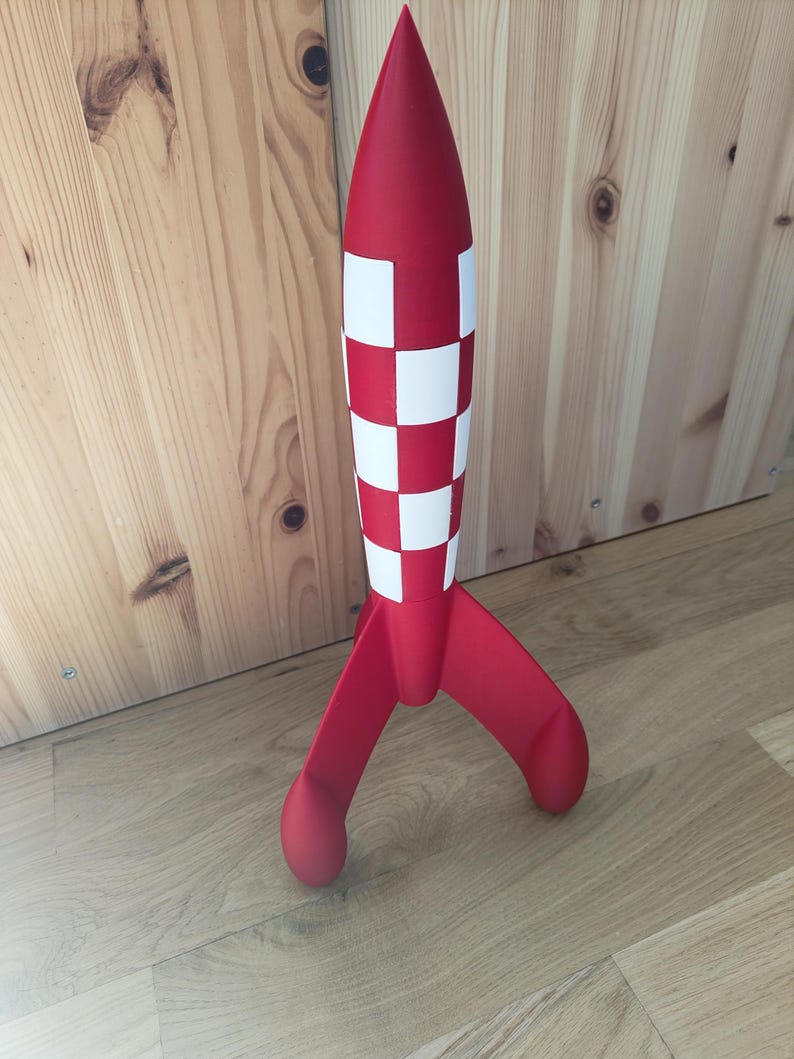 May include: A red and white rocket model with a checkered pattern on the body and three curved fins. The rocket has a pointed top and is standing on a wooden surface. A decorative item.