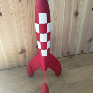 May include: A red and white toy rocket with a checkered pattern on the body and a rounded nose. The rocket has three fins and a red teardrop-shaped object suspended below. The toy is standing on a wooden floor.