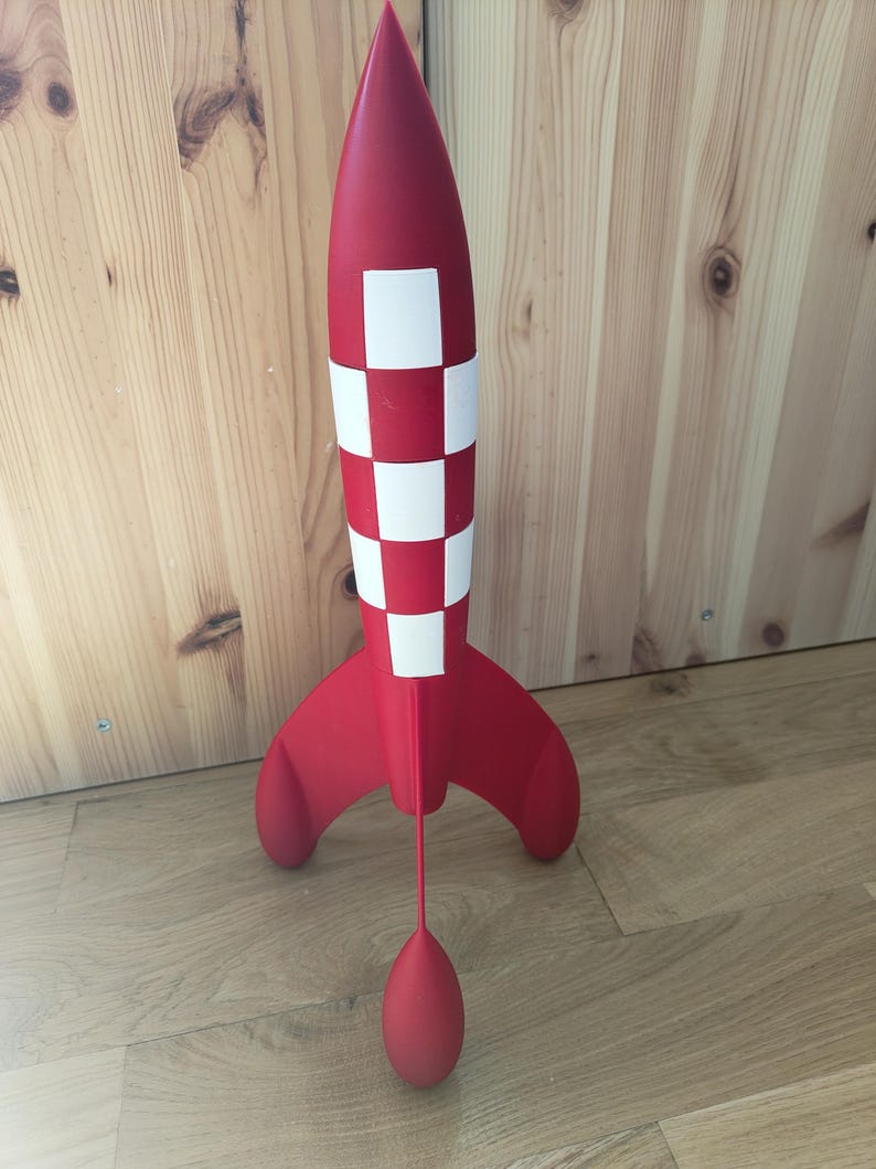 May include: A red and white toy rocket with a checkered pattern on the body and a rounded nose. The rocket has three fins and a red teardrop-shaped object suspended below. The toy is standing on a wooden floor.