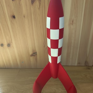 May include: A red and white rocket model with a checkered pattern on the body and three red fins. The rocket has a pointed nose and is standing upright on a wooden surface. A decorative item.