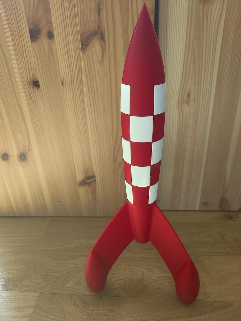 May include: A red and white rocket model with a checkered pattern on the body and three red fins. The rocket has a pointed nose and is standing upright on a wooden surface. A decorative item.