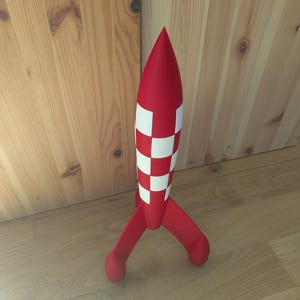 May include: A red toy rocket with white chequered detailing on the body and three red fins. The rocket stands on a wooden surface. The toy rocket is a children's toy.