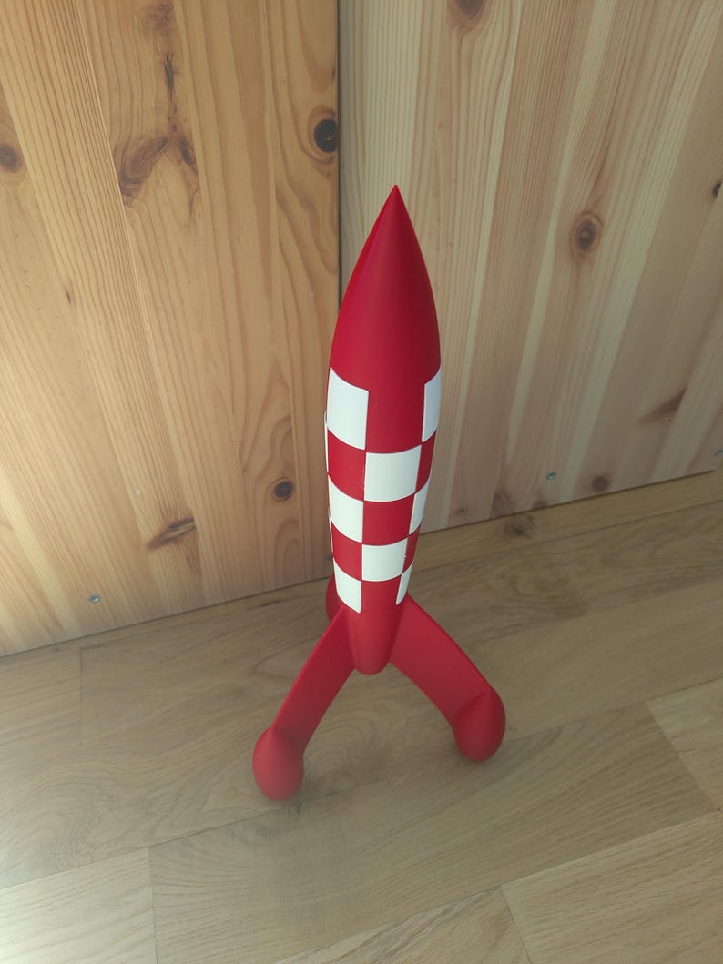 May include: A red toy rocket with white chequered detailing on the body and three red fins. The rocket stands on a wooden surface. The toy rocket is a children's toy.