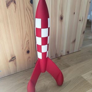 May include: A red and white rocket model with a chequered pattern on the body and three curved fins. The toy is standing upright on a wooden floor, with a wooden wall in the background.