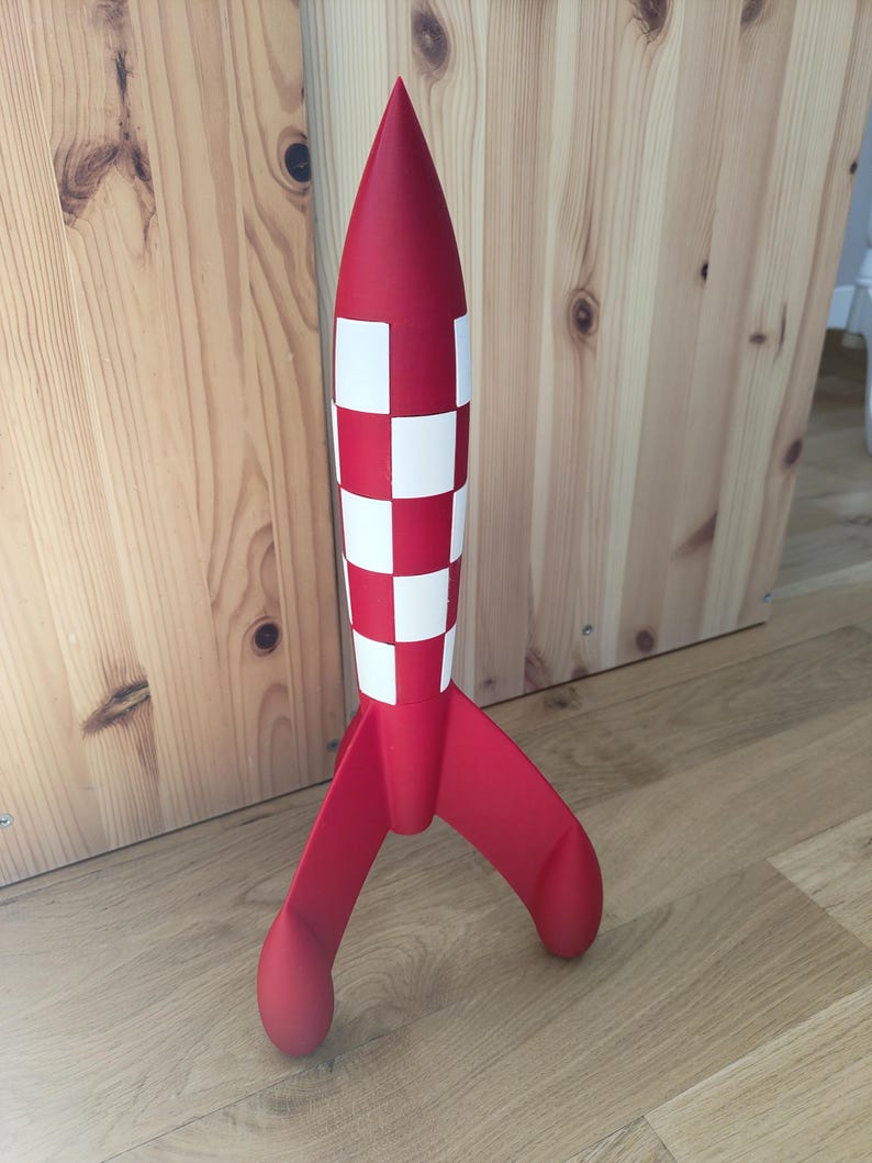 May include: A red and white rocket model with a chequered pattern on the body and three curved fins. The toy is standing upright on a wooden floor, with a wooden wall in the background.