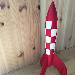 May include: A red toy rocket with white chequered accents. The rocket has three fins and a pointed nose. The rocket is approximately 46 cm tall and is made of a smooth material.
