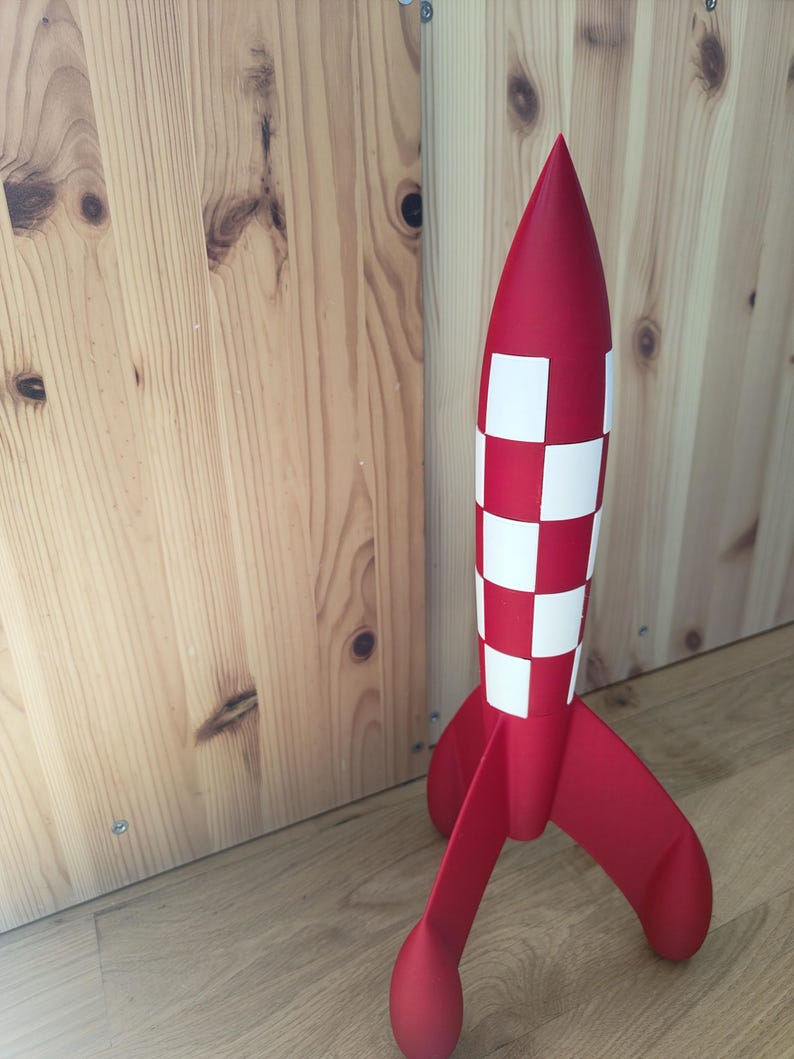 May include: A red toy rocket with white chequered accents. The rocket has three fins and a pointed nose. The rocket is approximately 46 cm tall and is made of a smooth material.