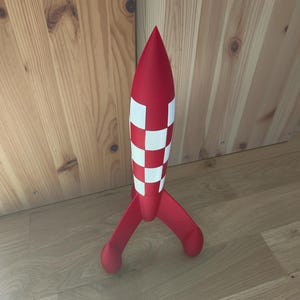 May include: A red toy rocket with white chequered accents and three rounded legs. The rocket has a pointed top and stands upright on a wooden floor. The rocket is a fun toy for children.