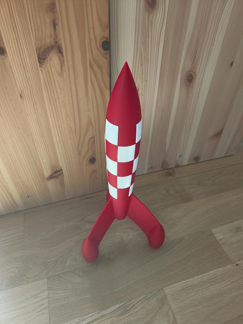 May include: A red toy rocket with white chequered accents and three rounded legs. The rocket has a pointed top and stands upright on a wooden floor. The rocket is a fun toy for children.