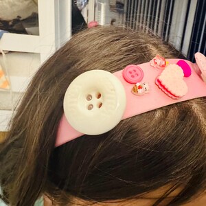 May include: A pink headband adorned with a large white button, pink buttons, and felt heart and bunny decorations. The headband is a playful accessory, perfect for adding a touch of whimsy to any outfit.