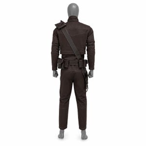 May include: Brown costume set on a mannequin. The outfit includes a jacket with shoulder strap, pants, and a belt with pouches. The jacket has a high collar and stitched details. The pants are straight-legged.