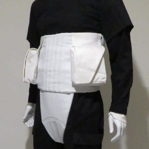 Op de afbeelding: Zwart-wit kostuum op een mannequin. De outfit bestaat uit een zwart shirt met lange mouwen, een zwarte broek en een wit vest met zakken. Witte handschoenen en ondergoed zijn ook zichtbaar.