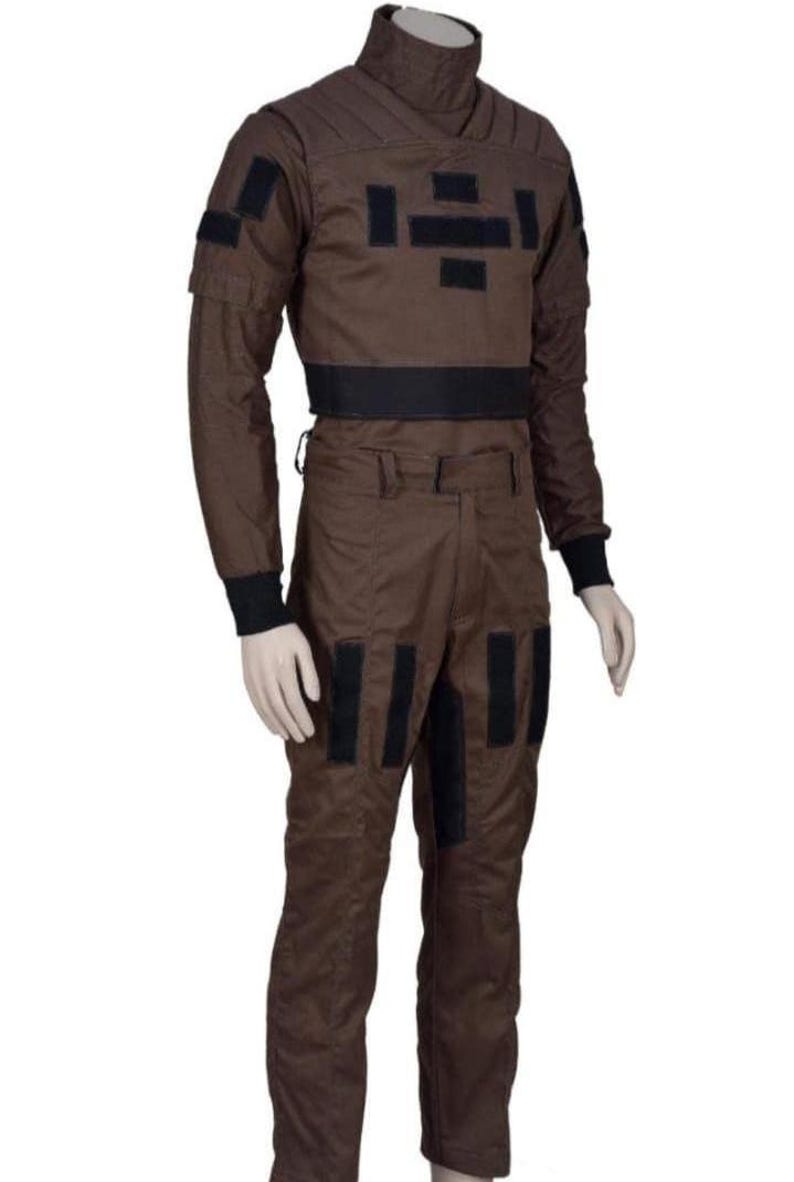 May include: Brown jumpsuit with black accents. The outfit features a high collar, long sleeves, and multiple black rectangular patches. A black band defines the waist. The pants have black patches on the legs. The jumpsuit is displayed on a mannequin.