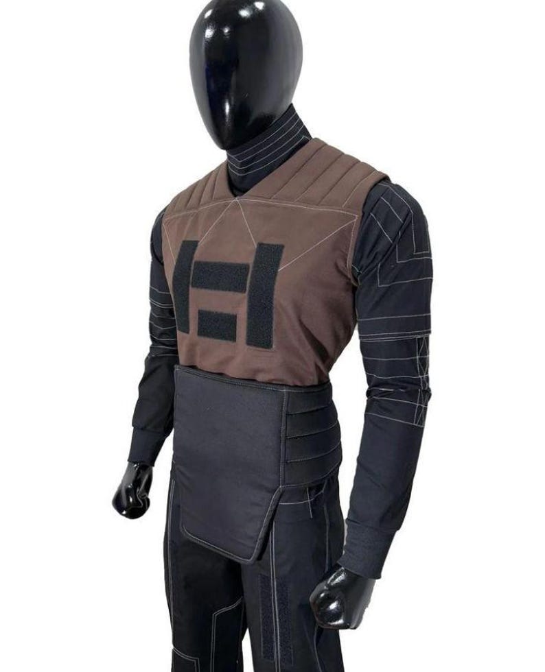 May include: A black long-sleeved shirt with white pinstripes worn under a brown quilted vest featuring a black rectangular patch design. A black padded waist belt is worn over black trousers with white stitching details on a mannequin.