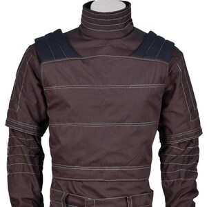 Op de afbeelding: Een donkerbruin shirt met lange mouwen, een hoge opstaande kraag, horizontale witte stiksels over de borst en mouwen, en donkerblauwe gewatteerde schouderpanelen. Het kledingstuk heeft een gestructureerd ontwerp met gelaagde details.