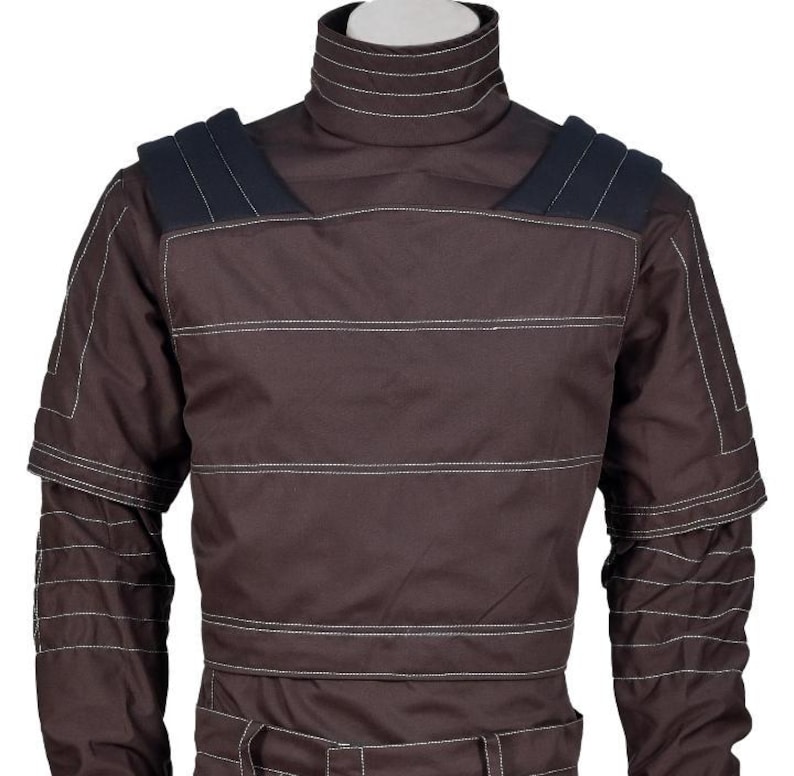 Op de afbeelding: Een donkerbruin shirt met lange mouwen, een hoge opstaande kraag, horizontale witte stiksels over de borst en mouwen, en donkerblauwe gewatteerde schouderpanelen. Het kledingstuk heeft een gestructureerd ontwerp met gelaagde details.