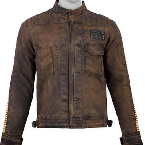 May include: Brown jacket with a stand-up collar, two chest pockets, and a small patch on the upper right chest. The jacket has a weathered appearance, with vertical stitching on the shoulders and a series of rectangular accents on the sleeves.