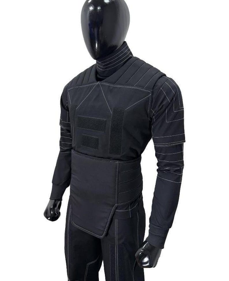 May include: Black protective suit with white stitching details, displayed on a mannequin. The suit features a high collar, quilted panels, and multiple Velcro closures. The long-sleeved top and pants are designed for full body coverage.