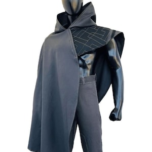 May include: A dark gray hooded cape and matching pants displayed on a black mannequin. The cape features a geometric pattern on the shoulder and a flowing design. The pants have a classic, tailored fit.