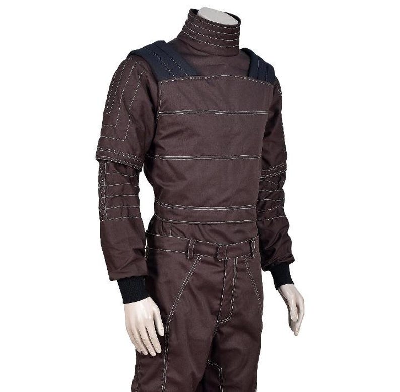 Op de afbeelding: Een donkerbruine jumpsuit met lange mouwen, een hoge opstaande kraag, zwarte schouderpanelen en zwarte geribbelde boorden. Het kledingstuk is voorzien van opvallende horizontale en verticale witte stiksels over het lijfje en de mouwen.